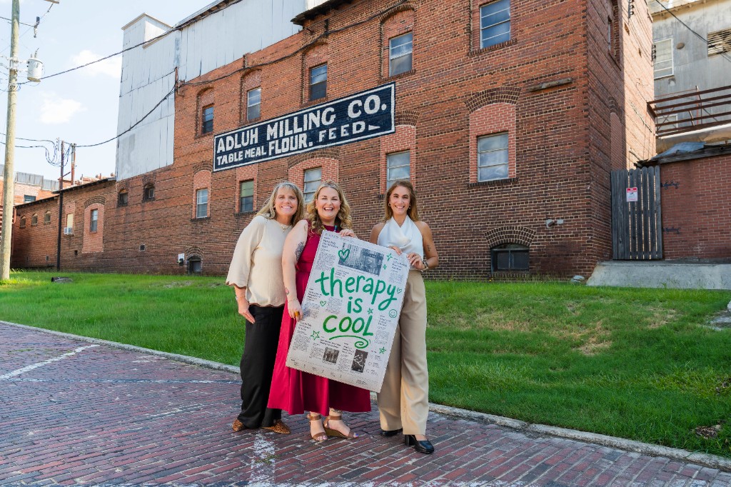 Jill Smith and two colleagues holding a Therapy is Cool sign in front of the Adluh Milling Co. building